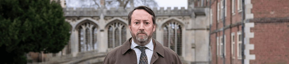 David Mitchell as John in Ludwig stares directly at the camera in front of a lovely Cambridge background 