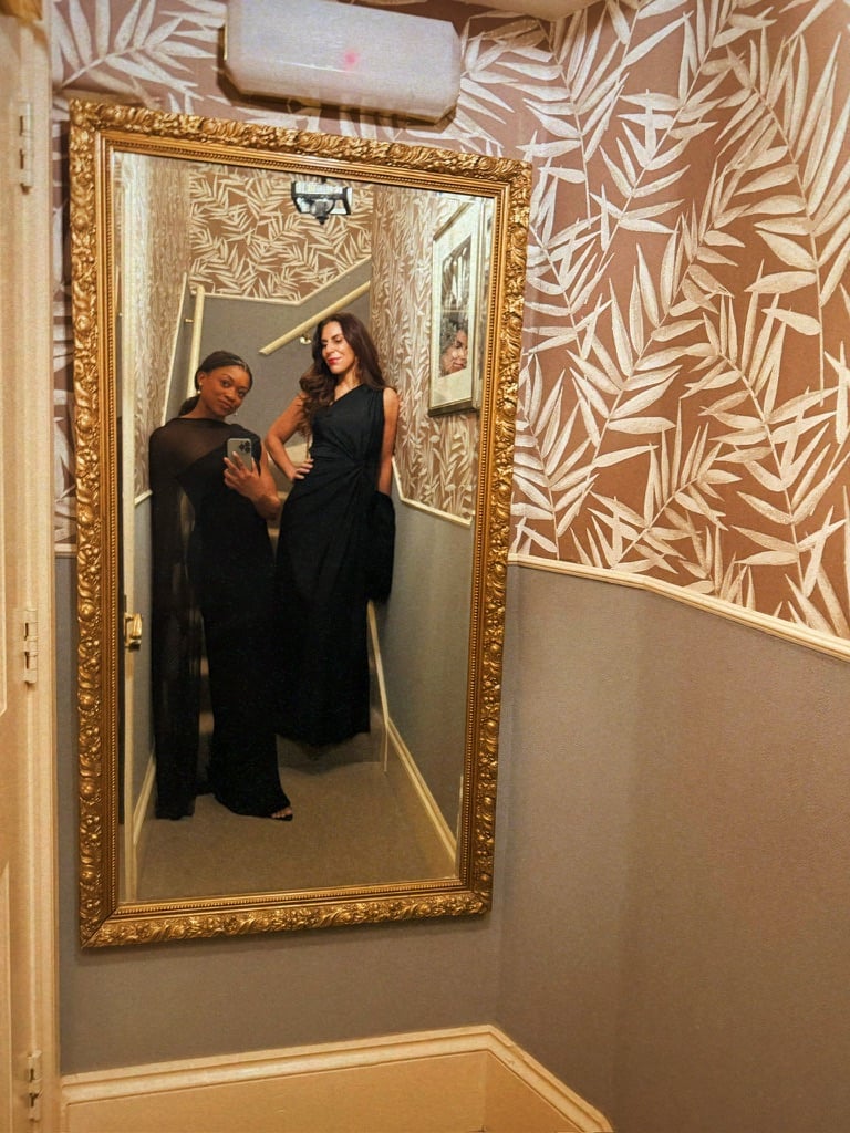 I’m standing in front of a large gold-framed mirror in a narrow hallway with leaf-patterned wallpaper, taking a mirror selfie. I’m wearing a long black evening dress with sheer detailing and holding my phone, while a friend beside me poses in a sleeveless black dress.
