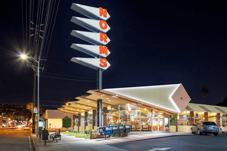 A photograph of a 1960 style diner brightly lit up at night. Its design is marked by large triangles -- there are a line of large and long horizontal triangles comprising the restaurant's roof, and the restaurant sign is five similarly long horizontal triangles stacked up on a pole, each one with a letter that together spell the name NORMS. The sky is completely dark and the restaurant shines in the darkness; the effect is very dramatic. On the far left you can see the suburban road on which the restaurant is located. On the far right a few cars are parked in the parking spots.