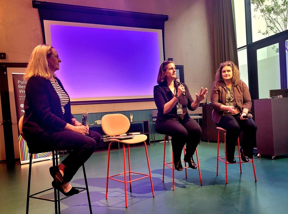 Three women sit on tall bar stools in front of a screen. The woman in the middle is speaking into a microphone.