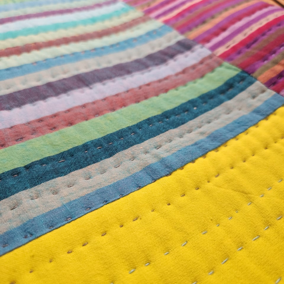 Close up photo of the back of String Quilt showing striped fabrics in shades of blue and purple, and a section of bright chartreuse. Sarah’s hand stitches are visible.