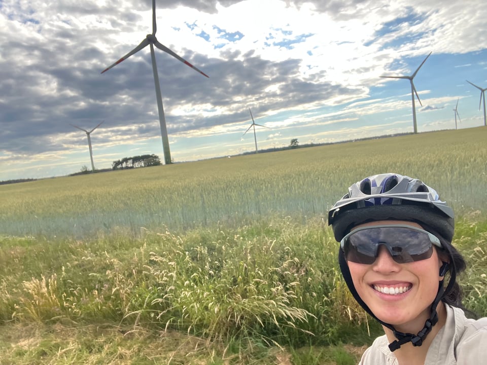 Jenny with helmet and cycling sunnies, doing a selfie against a backdrop of wheat and wind turbines.