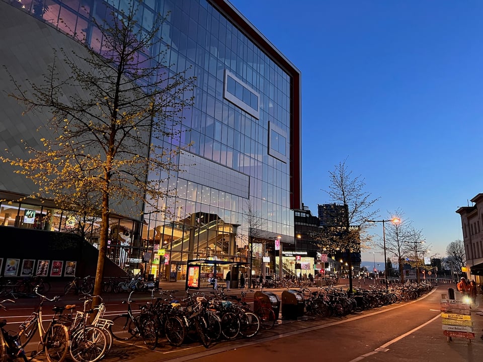 A photo of TivoliVredenburg in Utrecht, a large glass-fronted building facing an evening street with lots of bikes in front.