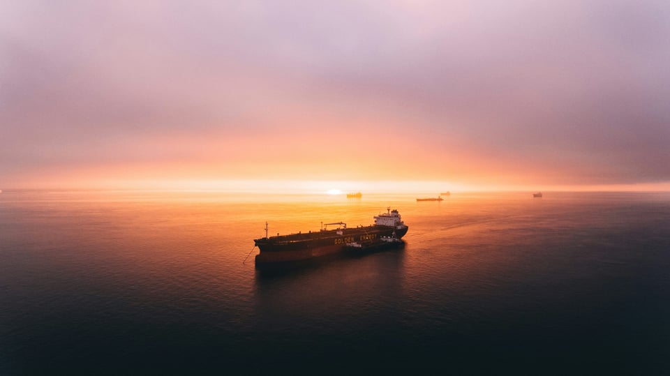 A tanker, anchored in the foreground, with more ships anchored in the background, closer to the horizon. The water is dark in the foreground, but takes on the reflection of the sunset further back in the photo to the point where the horizon is only discernible because the sun is visible halfway set below it. At the top are blue and gray from the clouds.
