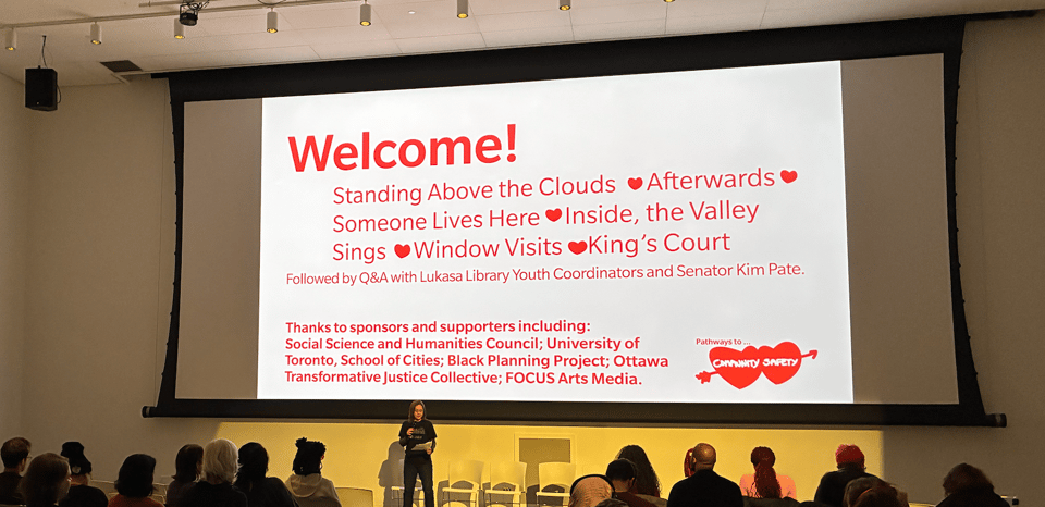 on a large film screen in red text with a pink background, Welcome! and the names of films: Standing Above the Clouds, Afterwards, Someone Lives Here, Inside, the Valley Sings, Window Visits, King's Court.<p>Further text: Followed by Q+A with Lukasa Library Youth Coordinators and Senator Kim Pate, Thanks to sponsors and supports.</p>
<p>A person stands welcoming the crowd who can be seen seated in front of the screen." class="" draggable="false"><figcaption><a target=