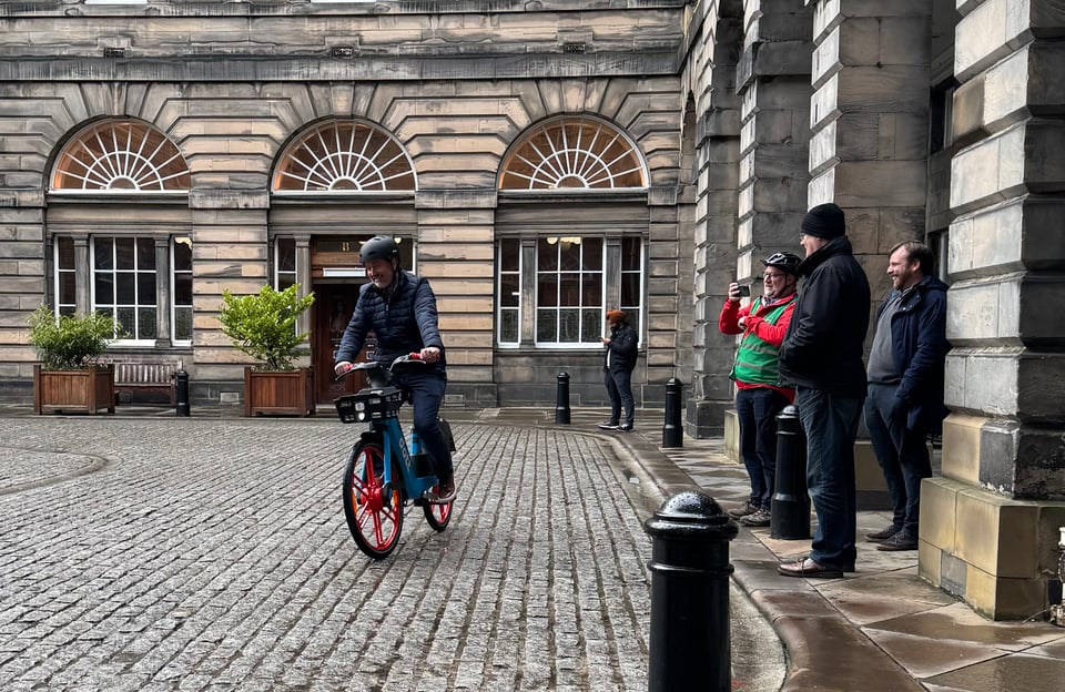 A man rides a brightly coloured ebike across grey setts - flat-topped cobble-like stones adorning the historic courtyard of the Edinburgh City Chambers as three other people look on with a smile.