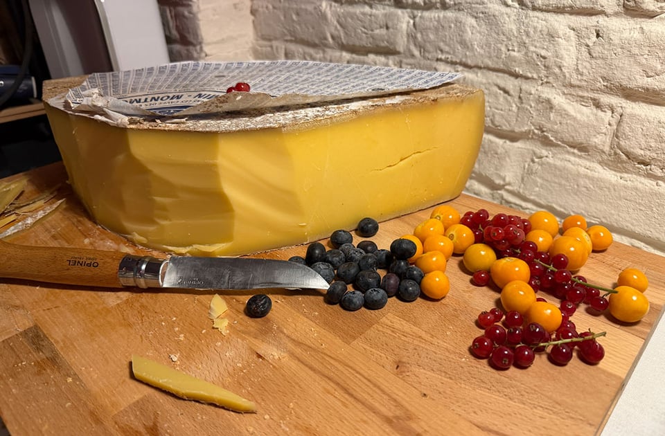 A large block of cheese sits on a cutting board next to fruits and a large knife.