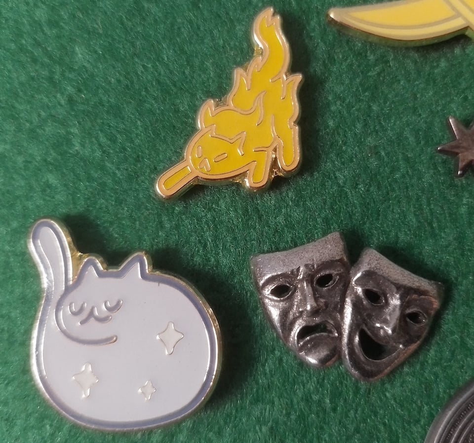 Three metallic pins pinned to a green felt board: a hissing cat spirit made of fire, a contented cat spirit made of stars, and a pair of Greek theatre masks