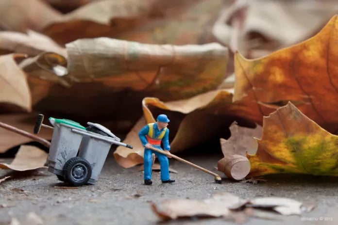 Photo of a tiny street cleaner with a tiny gray wheeled bin, pushing a tiny broom to shove enormous dead leaves out of the way on a sidewalk.