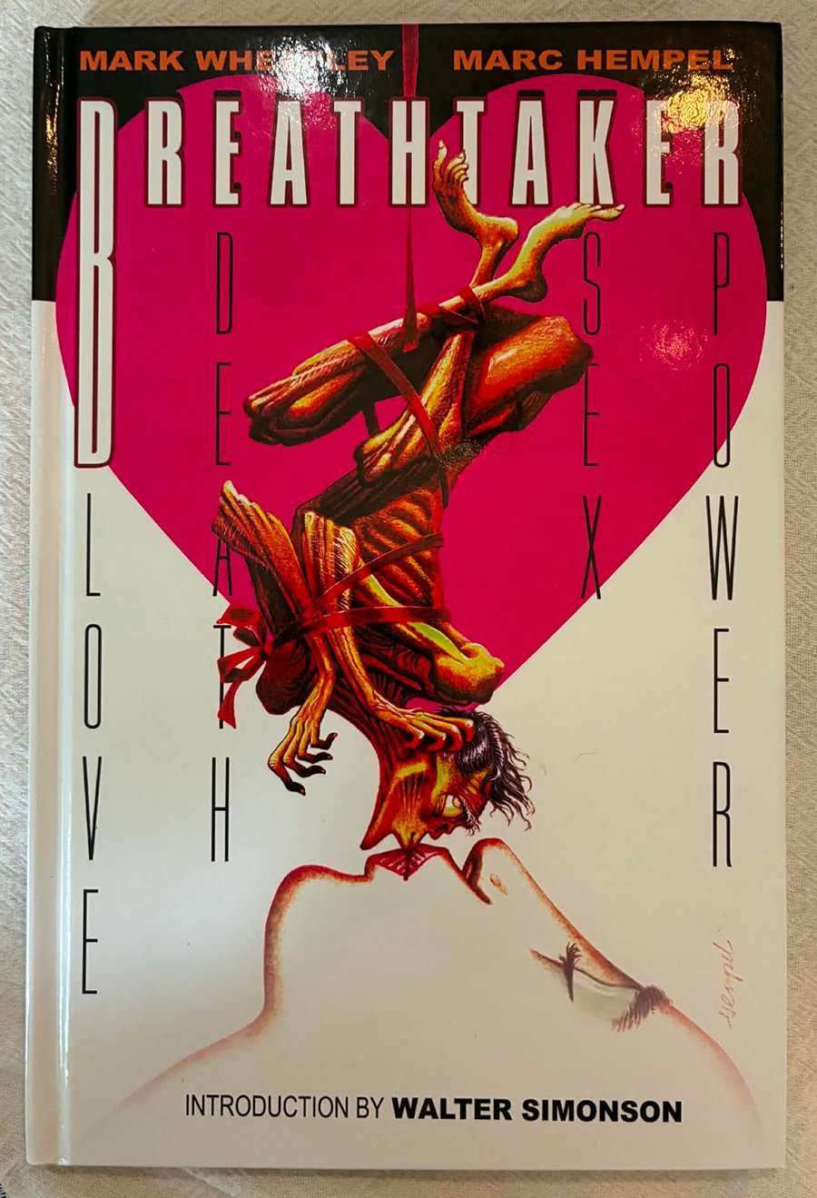 The hardcover volume Breathtaker sitting on a tablecloth. Cover shows an emaciated man tangled up and dangling over a woman's face, she is kissing him.