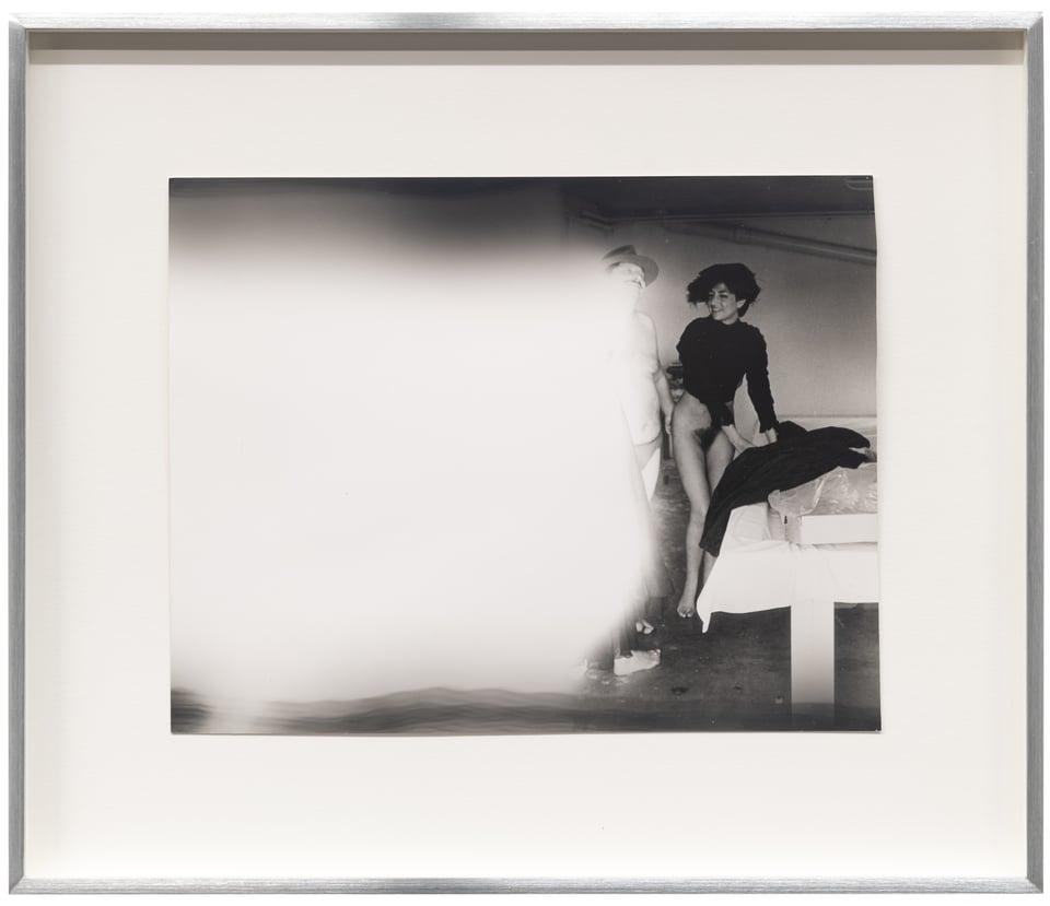 In this black-and-white print, artist Alix Cléo Roubaud has, though the darkroom process of "dodging" has obscured more than half of the image with a white cloud. You can see a white woman with dark hair, nude from the waist down, laughing/smiling at a man in a hat and underpants who is only semi visible. they are in a room, on the right side.