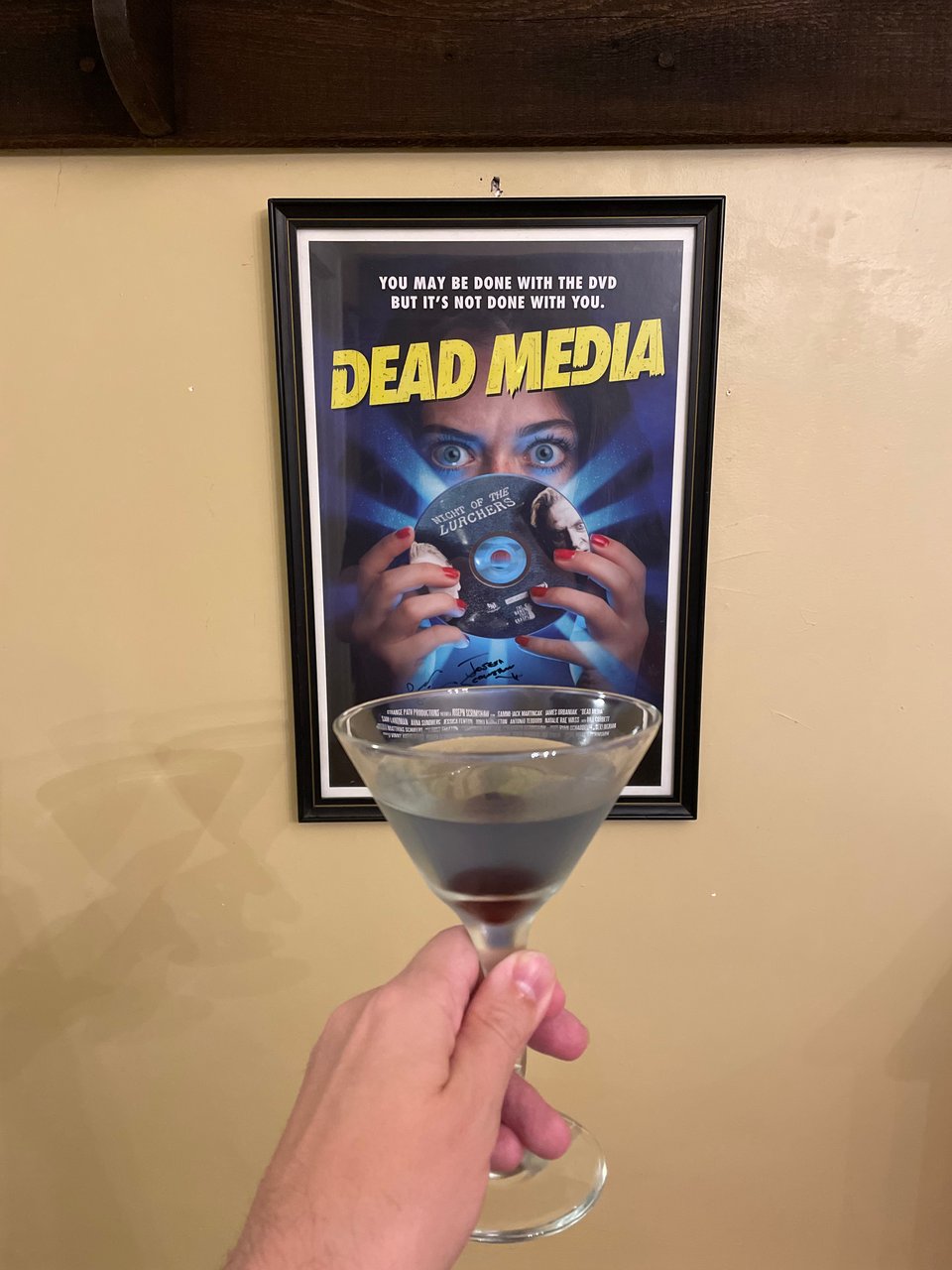 A man holds a cocktail up to a poster for the horror movie, Dead Media