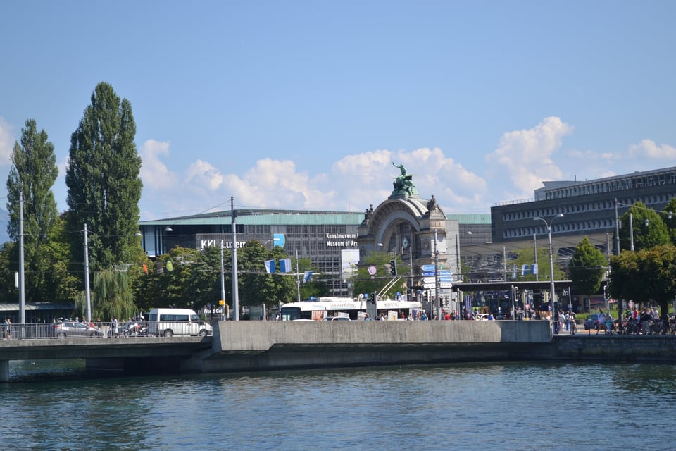 A very strange sculpture on the far side of water, with a tram behind it and a train station in the background.