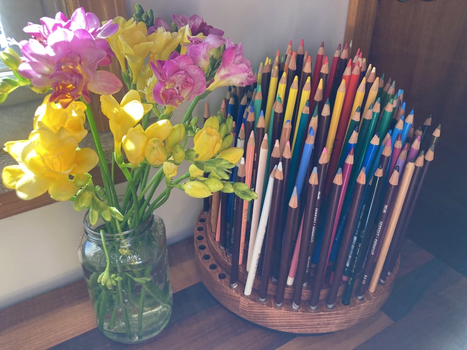 A round, wooden pencil organizer on a brown wooden desk next to a bunch of pink and yellow freesia flowers in a glass mason jar. The organizer is filled with colored pencils from various brands in rainbow order.