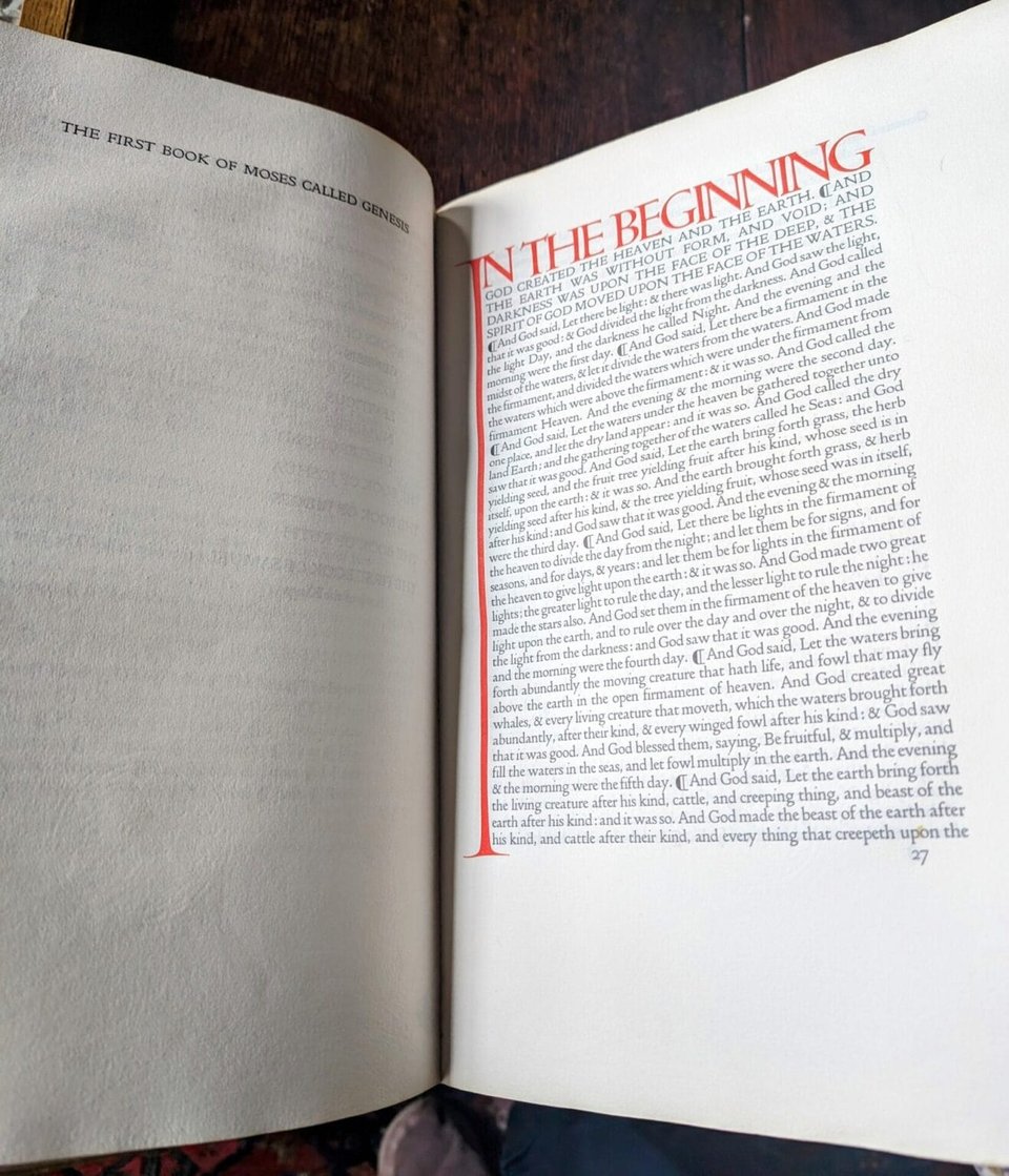 A photo of the opening passage of the Bible in a very large book. The top line is large, bold, uppercase letters, saying "in the beginning", with the eye stretching downwards, the entire length vertically of the page of text. The rest of the text is small and normal sized and in black.