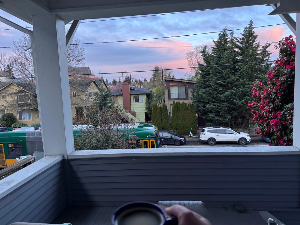 A view from the front door: a bus passes by, a sunrise paints the sky pastel blues and pinks, and Maria's hand holds a cup of coffee in the foreground.