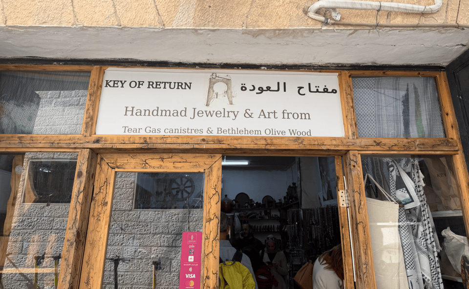 Photograph of a storefront with a sign that says ‘Key of Return’, along with a drawing of the sculpture of the entrance with the sculpture of the key, shown in the first photo above.  The sign has Arabic writing, and says also:  Handmade Jewelry & Art from Tear Gas canistres & Bethlehem Olive Wood. The sign is above wooden-framed glass doors, with interior displaying various art pieces and black and white keffiyehs inside
