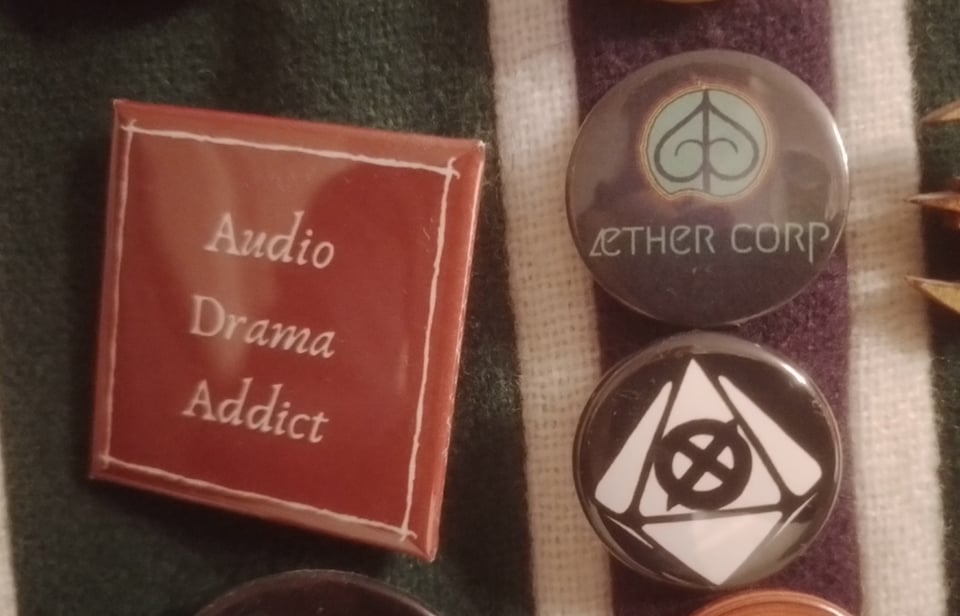 Three badges pinned to a striped scarf. A big square red one that says Audio Drama Addict in a script font, a small round black one that says Aether Corp under a teal logo a bit like a spade from a deck of cards, and another small round black one with a stylised illustration of a ten sided die, with all the faces blank except for one with an X in a circle on it.