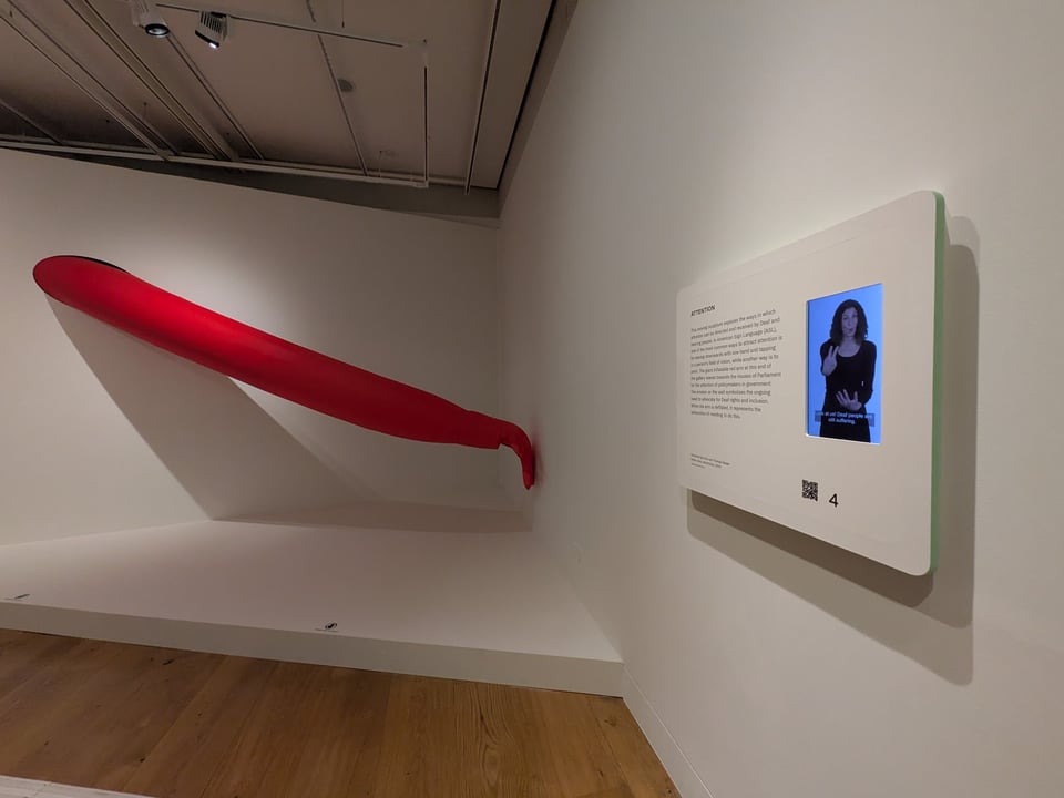 Artwork in 1880 THAT: A massive inflatable red arm is waving in the ASL sign for "attention." On the wall, a label is presented in both English and BSL (with captions).