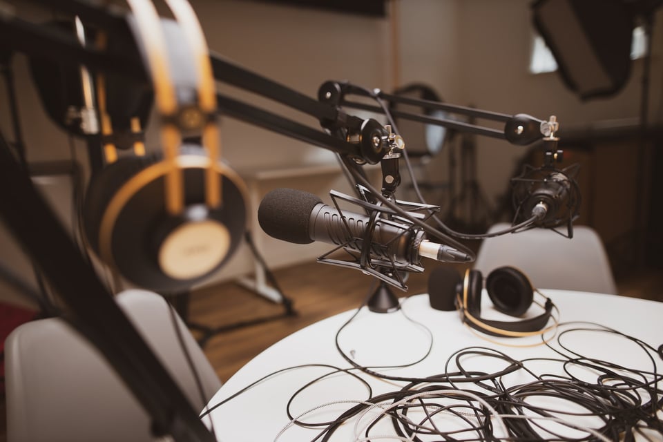 A podcast studio. A microphone on a boom is in focus. A set of headphones in the foreground, and the table in chairs in the background are out of focus. Photo by Jonathan Farber on Unsplash.