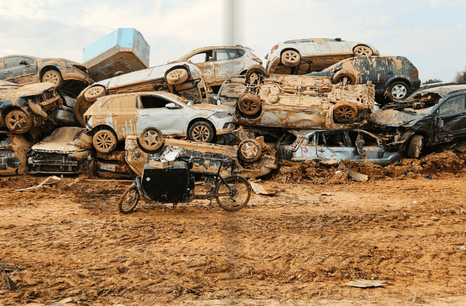 On a muddy street, a cargo bike awaits use in front of a pile of wrecked cars.