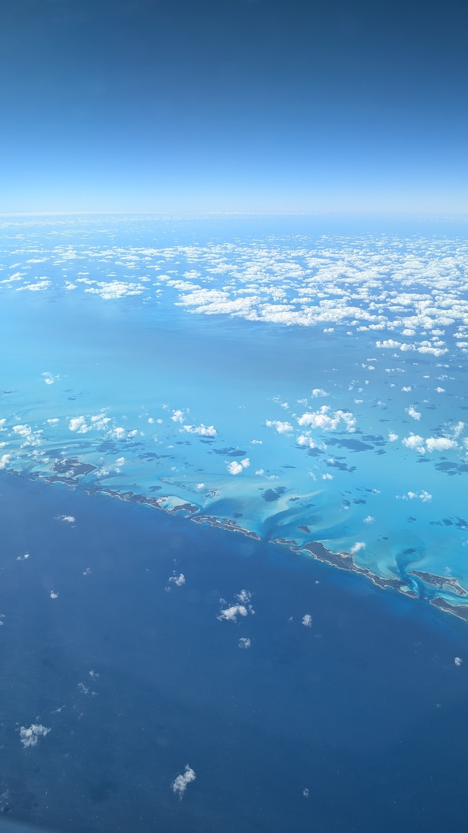A cell phone photo of beautiful blue sky with low-altitude white clouds skimming over an incredibly blue ocean, which is bordered with small green and brown islands. I took this photo on the flight from Florida to Barbados.