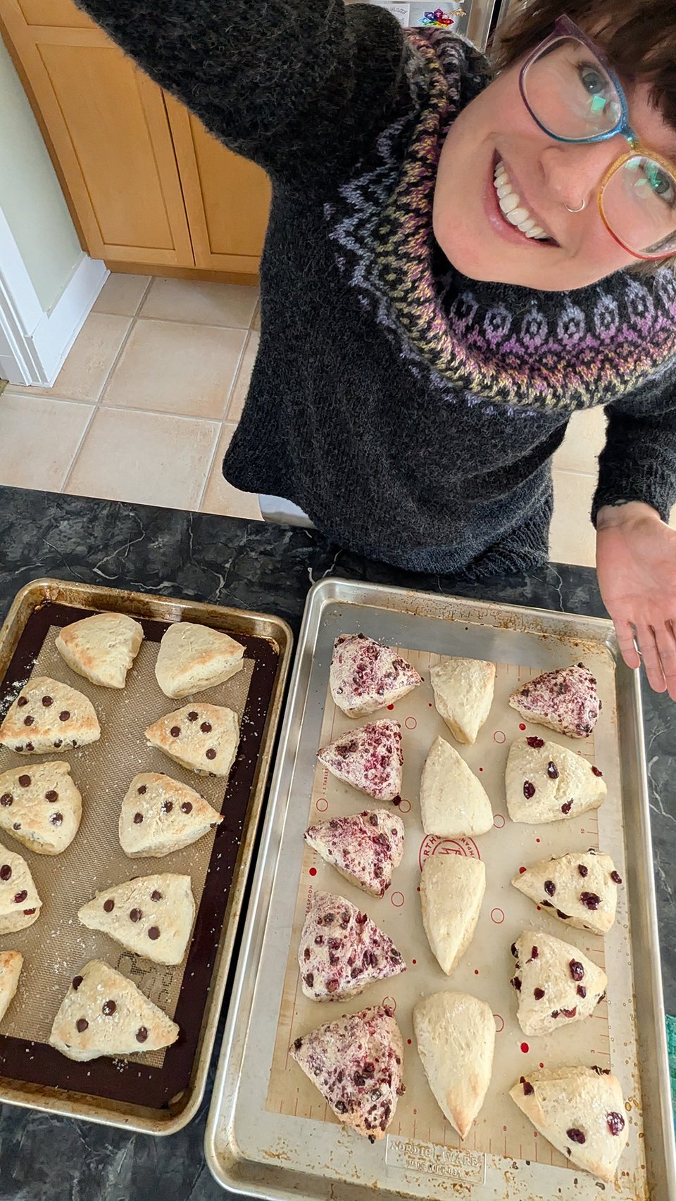a selfie taken from above of Maia showing off two baking trays full of triangular scones. There are four varieties and they all look extremely delicious (and they are.)