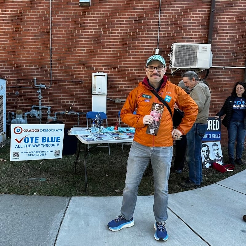 A white man with a mustache stands on a sidewalk in front of a folding table covered with campaign literature and sample ballots. He has a "Latinos for Harris Walz" sticker on one side of his jacket, and on the other an "I voted" sticker and a name tag reading "Cristóbal Palmer town council candidate"