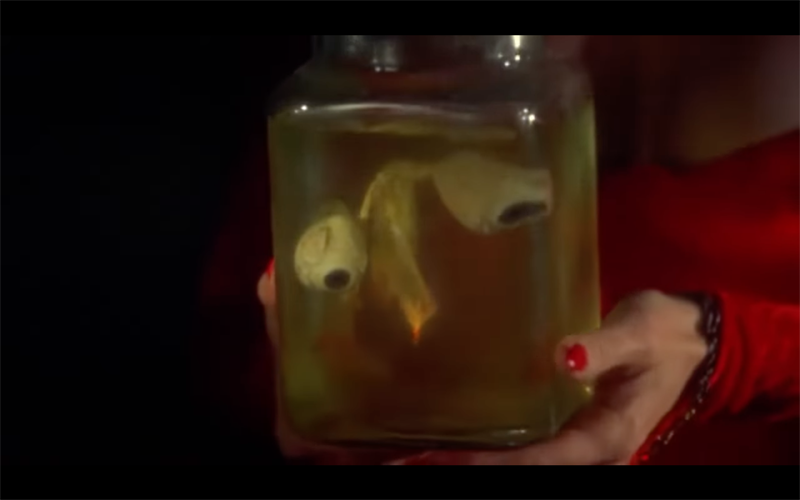 Close-up from Ruby (1977) of Piper Laurie's hands holding a jar with a pair of unconvincing white rubber eyes strung with red thread floating inside