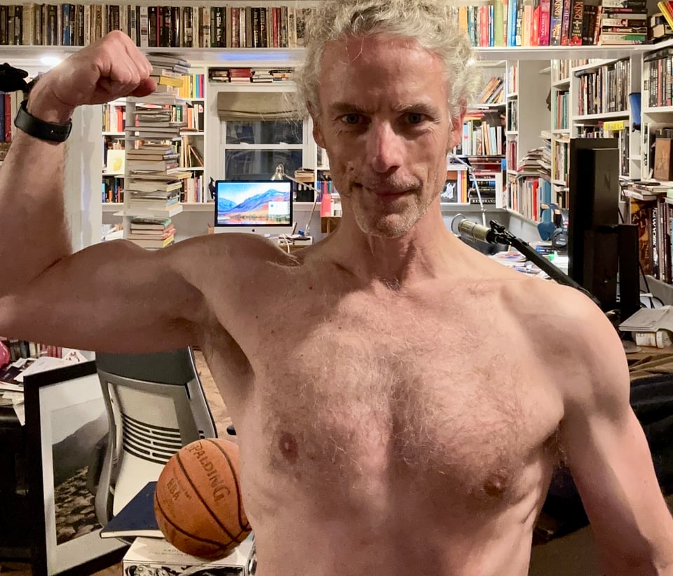 Photo of shirtless white man flexing his right bicep for the camera. There are a LOT of books in the background, as well as a basketball and some computers