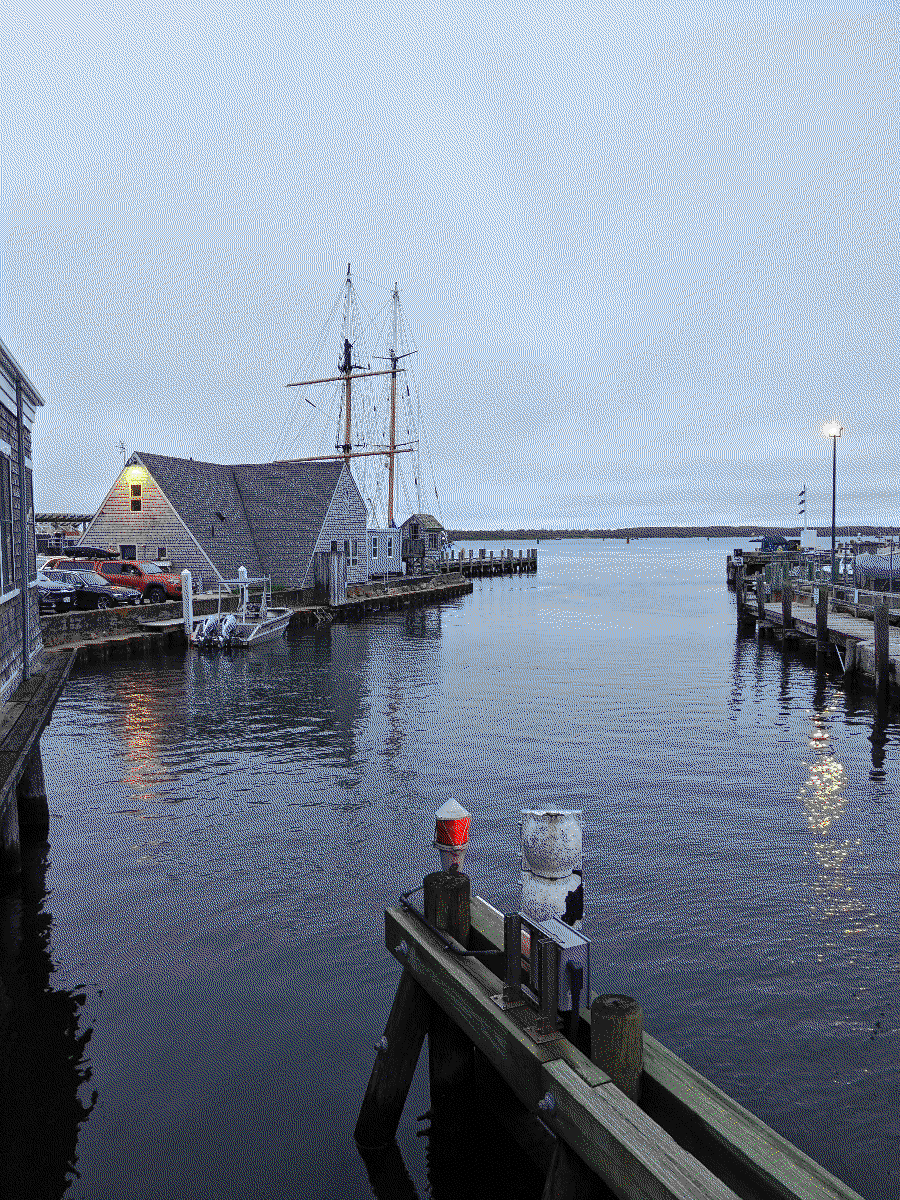 A very still, gray view of a quiet harbor. An old-looking red warning light faces out towards the ocean. A tall sailing vessel is docked in the distance.