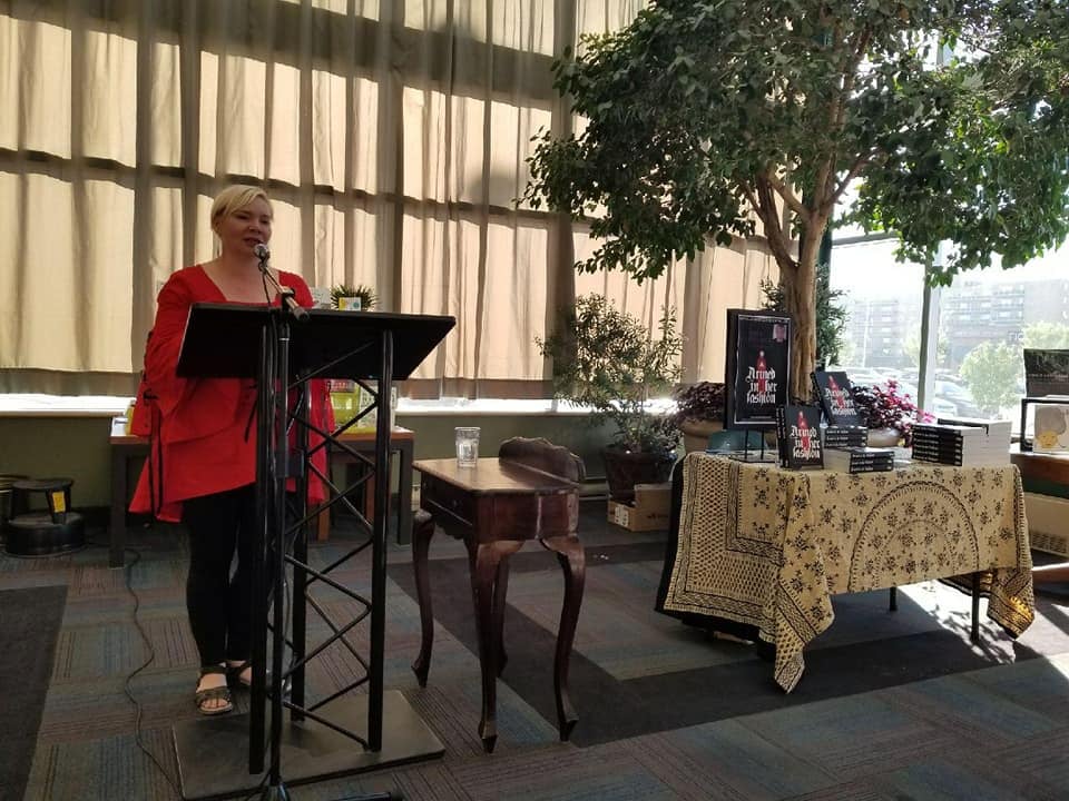Kate at a lectern with a red medieval tunic over leggings, and floppy assymettrical hair. There's a book table next to her with an actual tree behind it.