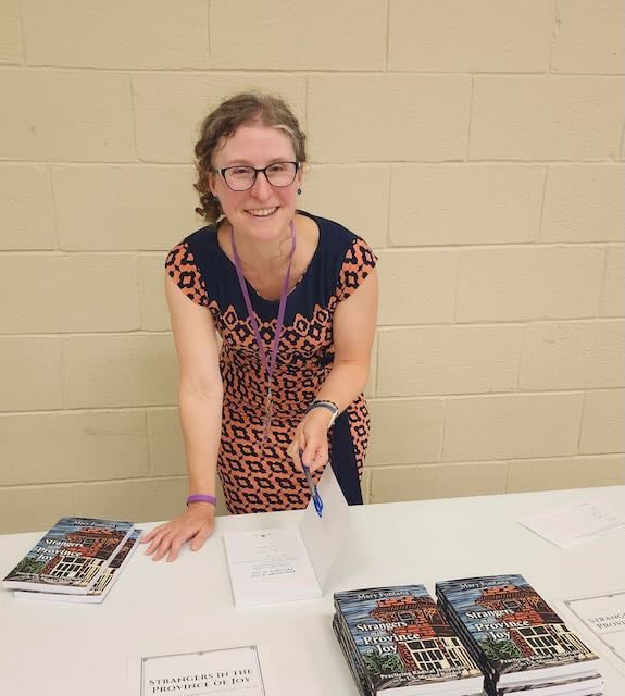 A smiling woman in a patterned dress is standing at a table with books and paper, holding a pen and preparing to write.