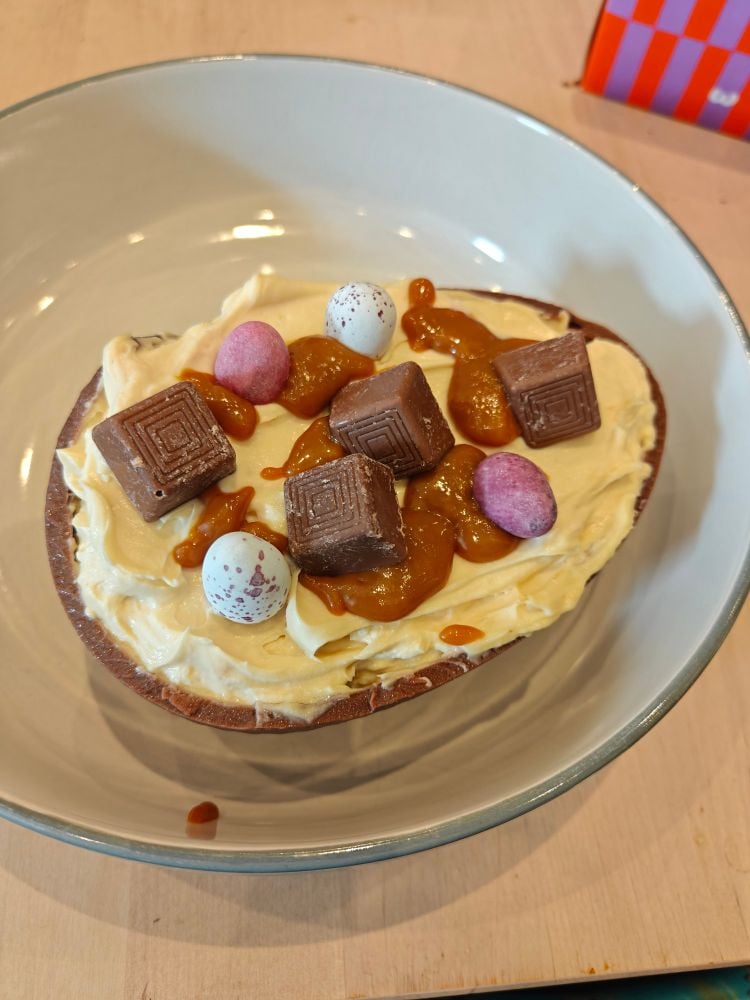 Half a chocolate easter egg filled with cheesecake. On top of the cheesecake is some caramel drizzle, mini eggs, and munchies