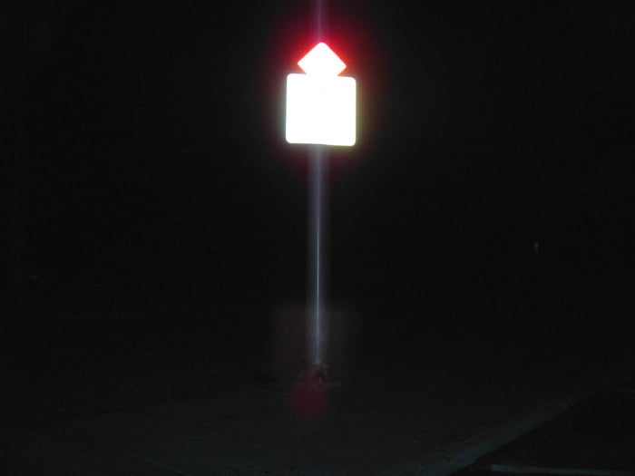 A photo of a reflective sign at night on a stout pole. It is unclear what the sign says, but there is a red diamond perched on a white square. The camera flash or nearby light is enough to overexpose the sign and pick out the shape of the pole in the dark but that is all.