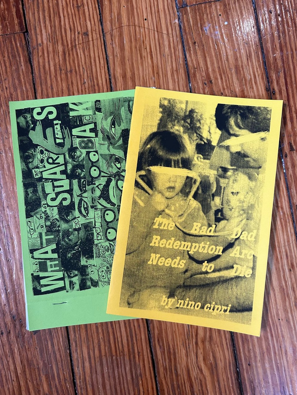 Two zines on a wooden floor. One has a bright green cover with a collage of eyes, and is titled "What Stares Back." The other has a neon yellow cover, depicting a child holding a toy stethoscope to her father's chest, with both their eyes scratched out, and is titled "The Bad Dad Redemption Arc Needs To Die."
