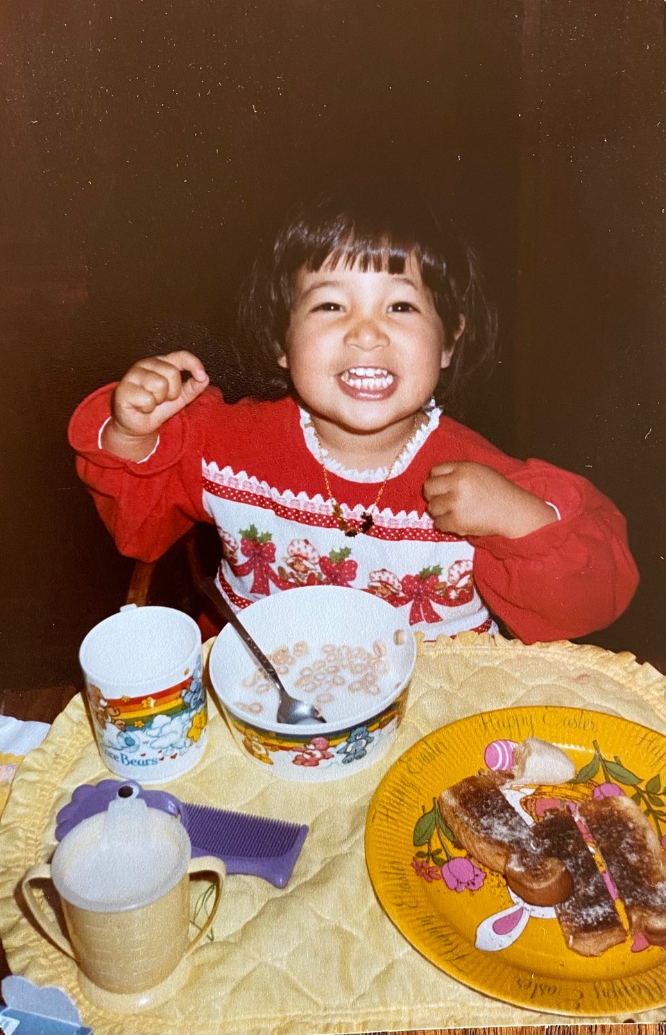 a young brown child smiles at the camera with food in her mouth in front of her is a bowl of cereal with milk, a spoon, two mugs and a plate with buttered toast