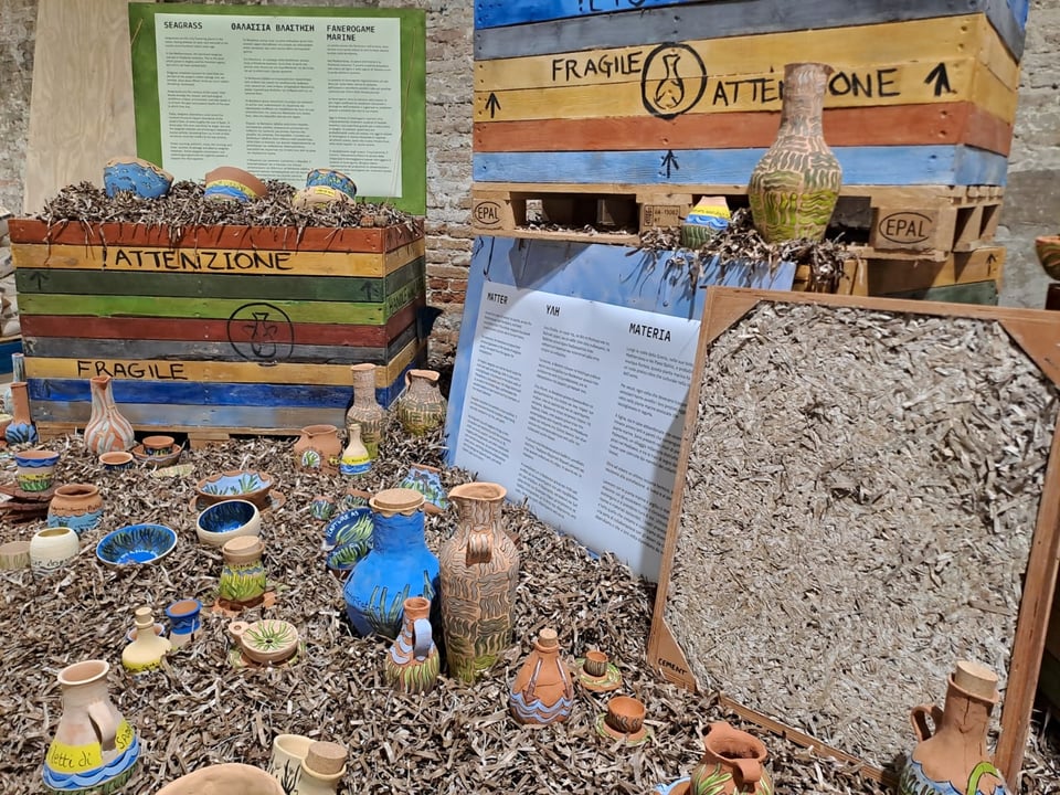 An image of Vessel's installation at the Venice Biennale: four brightly-coloured wooden crates are surrounded by piles of dried seagrass, in which are embedded over a hundred clay vases and other vessels, all painted with seagrass imagery and messages.
