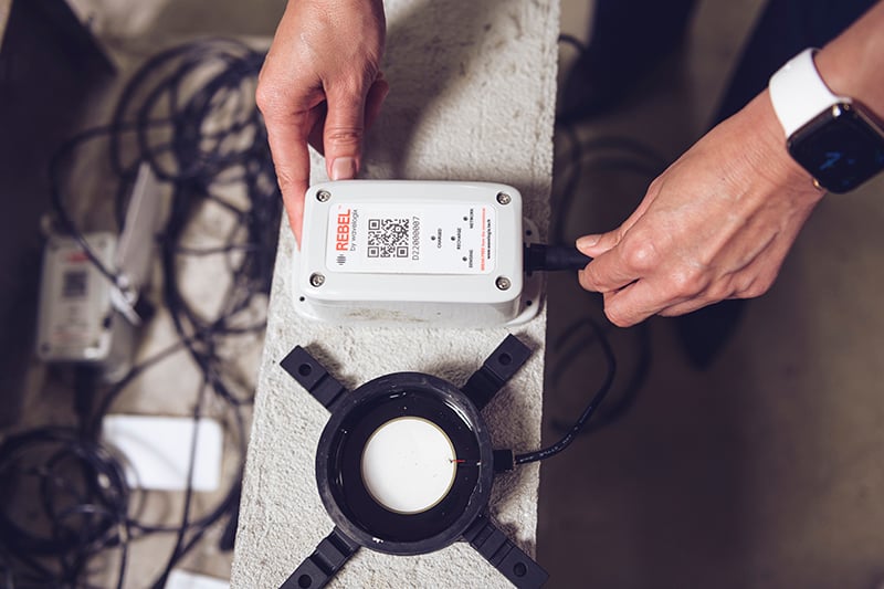 The REBEL Concrete Strength Sensing System, which is a circular sensor intended to be embedded into concrete, with wires to a read-out box that can be connected to by smartphone
