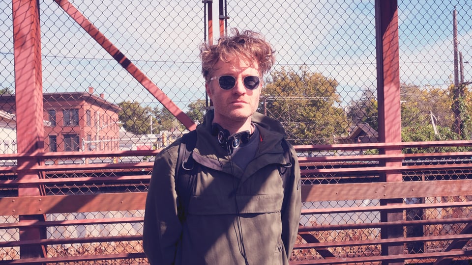 A man with a tangle of red hair wearing sunglasses stands on an iron bridge near downtown Northampton.