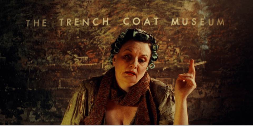 A still from The Trench Coat Museum music video with a woman in curlers, smoking.