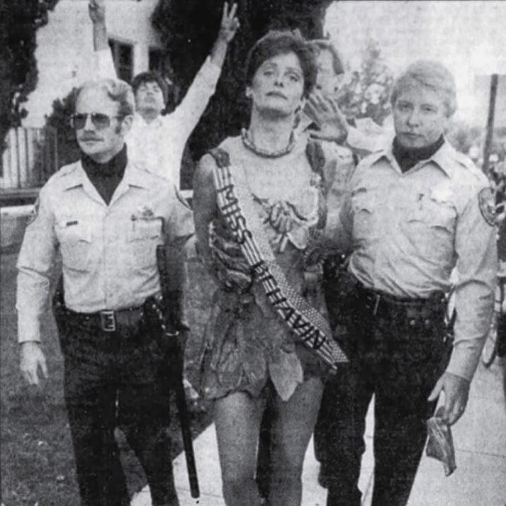 A black and white image of a woman being escorted by two cops, with her hands behind her back. She's wearing a dress made out of meat, and a sash that says "Miss Behavin"