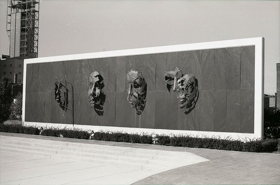 Black and white photo of four bronze masks, each only depicting a portion of a face. The sculpture is entitled, Faces of our Times, and resides at the Midland Insurance Plaza at the Midland Mutual Life Building.