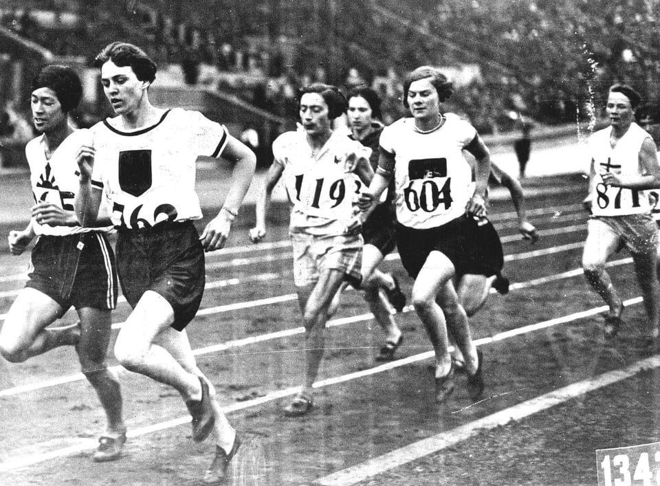 A black and white photograph from 1928 showing a pack of runners on a track, each wearing dark shorts and light tops with big numbers pinned to them