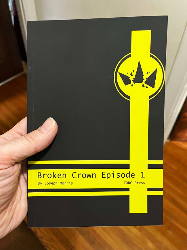 My hand holding a black paperback copy of Broken Crown episode 1