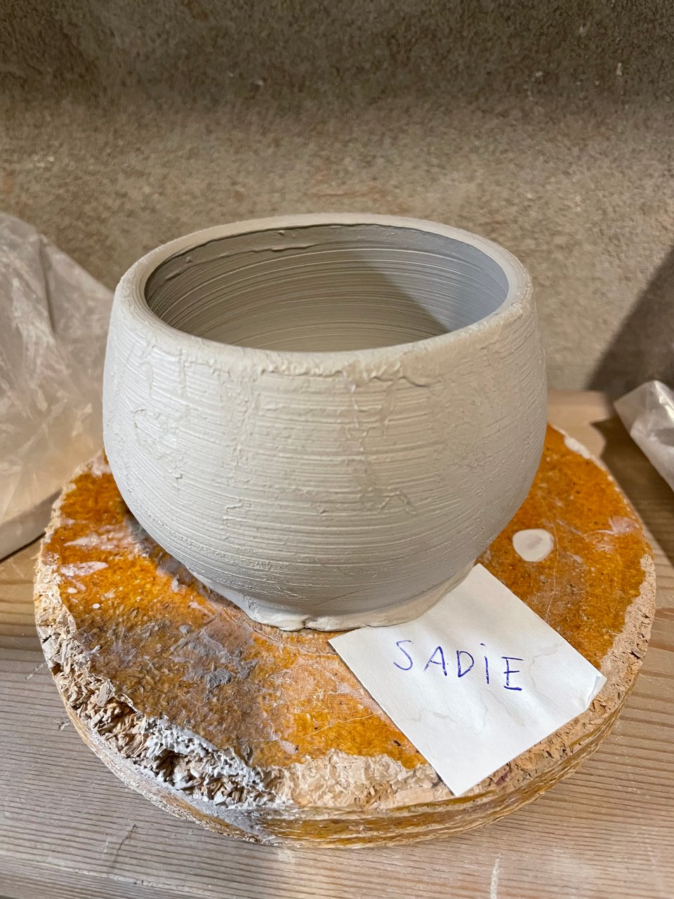 An unfinished, round piece of pottery sits atop a round piece of wood particle board. The pottery is dove grey with lines from the potter's hands. A small square white slip of paper on the board says S A D i E  in blue ball-point pen.  