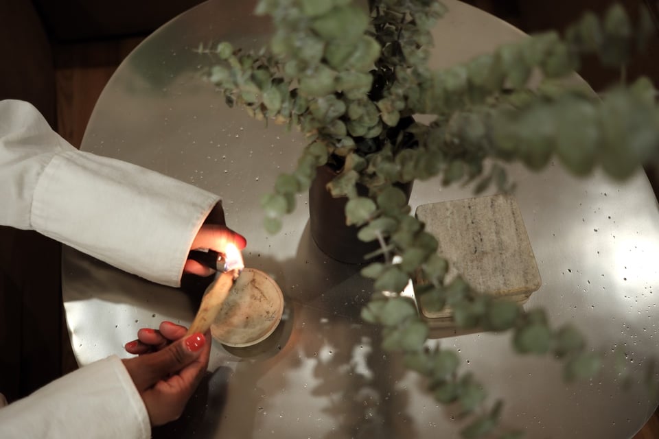 black hands with red nails hold a lighter to palo santo next to a vase of dried eucalyptus