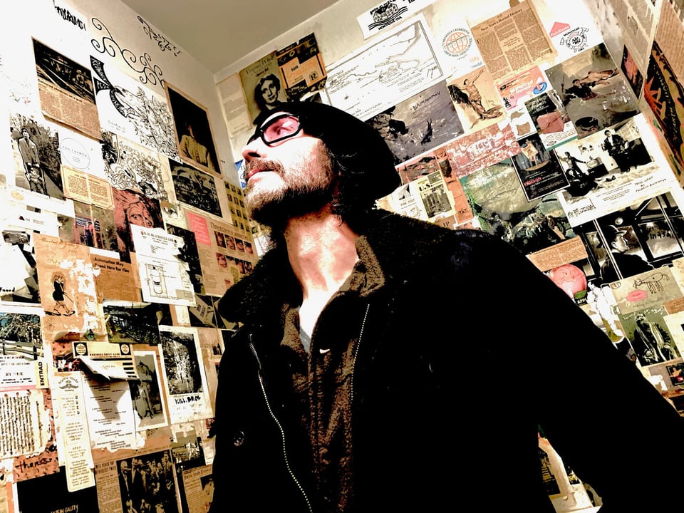 A man with a trim beard, tinted glasses and a winter hat looks up and to his right in a photo taken from below.