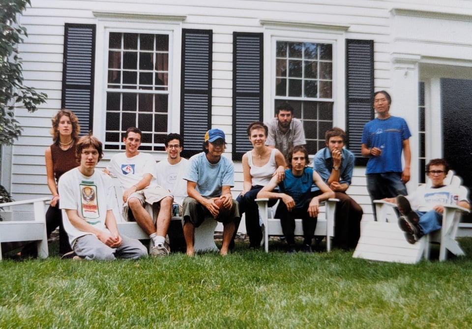 A group of 23 year olds sit in front of a white farm house. It's summer, and no one has showered.