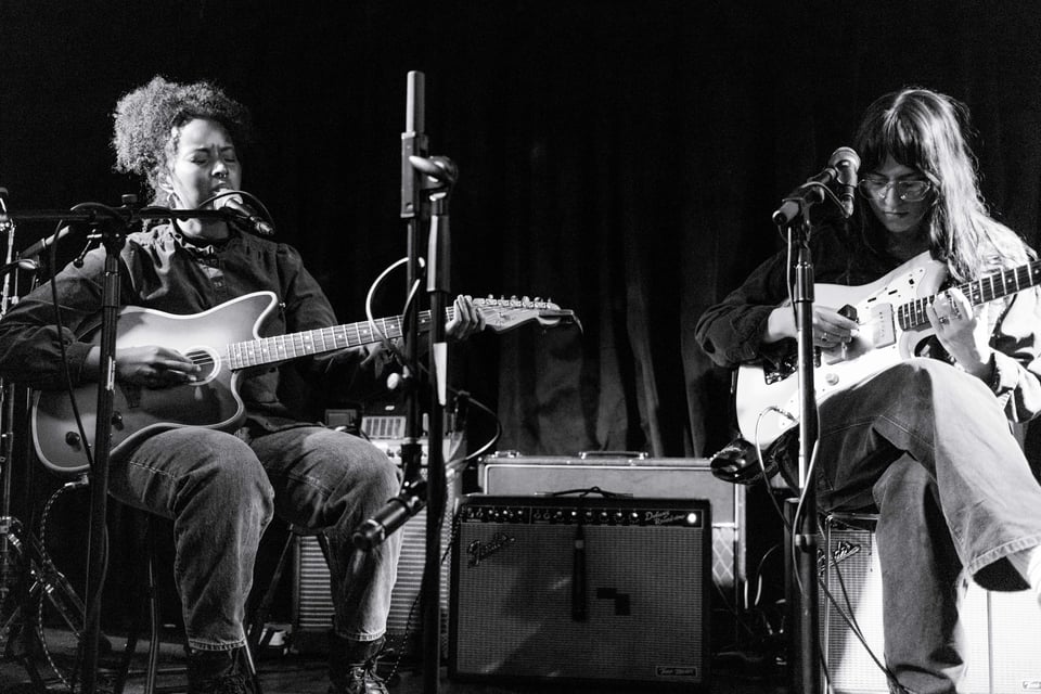 Maryam of poolblood is on the left, singing and playing guitar. Claire Ozmun is on the right of the photo, looking down at the guitar she's playing. black & white.